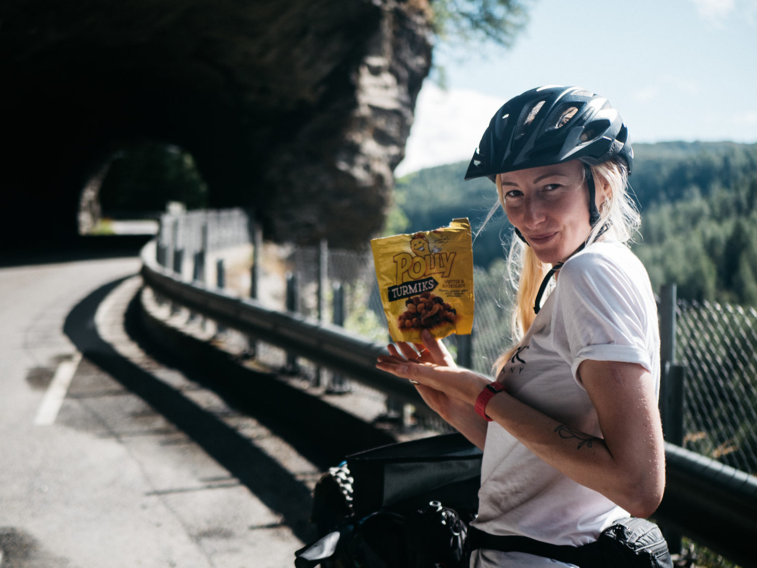 A cyclist pauses on a mountain road holding a bag of sweets, cliffs and hills behind